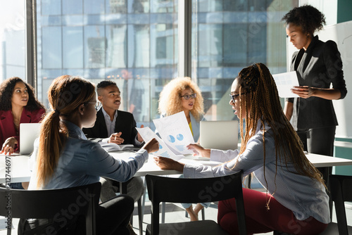 Black female project executive give printed handout materials to diverse teammates on meeting in office. Multiethnic staff group discuss sales result on briefing using paper reports with charts graphs photo
