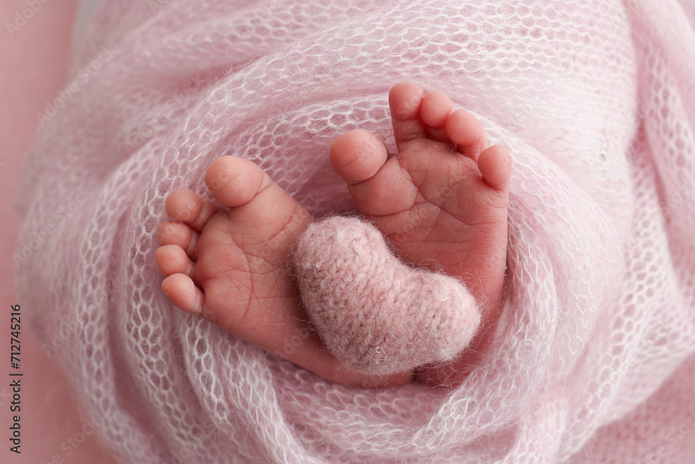 The tiny foot of a newborn baby. Soft feet of a new born in a pink wool blanket. Close up of toes, heels and feet of a newborn. Knitted white heart in the legs of a baby. Macro photography. The tiny foot of a newborn baby. Soft feet of a new born in a pink wool blanket. Close up of toes, heels and feet of a newborn. Knitted white heart in the legs of a baby. Macro photography.