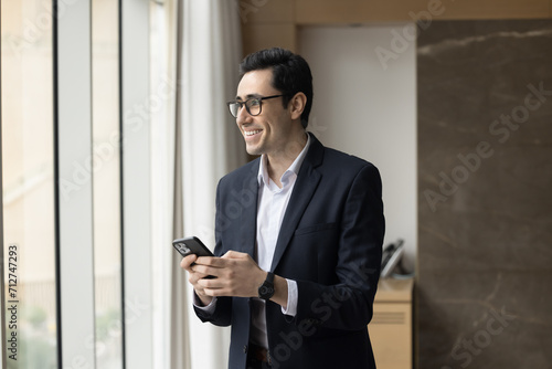 Cheerful young Arab entrepreneur man holding mobile phone, standing at office window, looking away, smiling, enjoying wireless online communication, thinking on job success