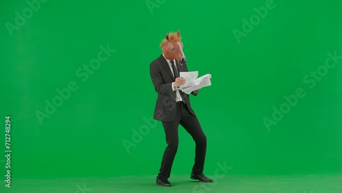 A man in a business suit with a horse head mask on a green studio background. A businessman is looking through documants and dancing merrily. Hard office work concept. photo