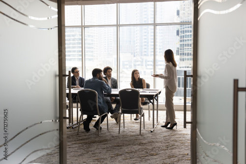 Candid shot through doorway of diverse business team and female leader discussing teamwork at large table. Group of young entrepreneurs negotiating, networking in modern office meeting room