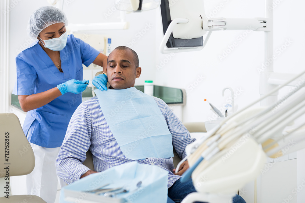 Obraz premium Portrait of adult hispanic man sitting on dental chair while woman dentist preparing him for medical examination in clinic
