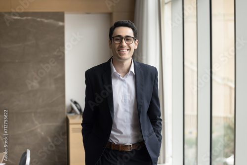 Happy young Arab company CEO man in formal shirt and glasses standing at workplace in office, keeping hands in pockets, looking at camera, smiling, laughing. Confident business professional portrait