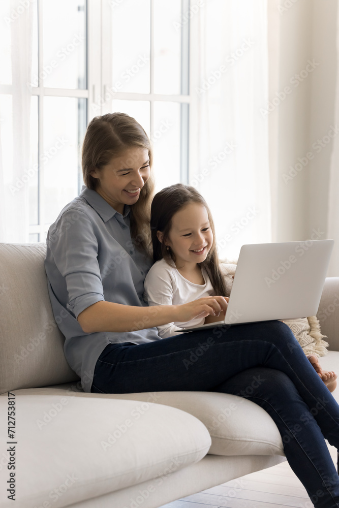 Caring attractive mother spend weekend time with cute little daughter at home, sit on sofa in living room, using laptop, shopping online, watch funny video on computer together, enjoy rest on internet Caring attractive mother spend weekend time with cute little daughter at home, sit on sofa in living room, using laptop, shopping online, watch funny video on computer together, enjoy rest on internet