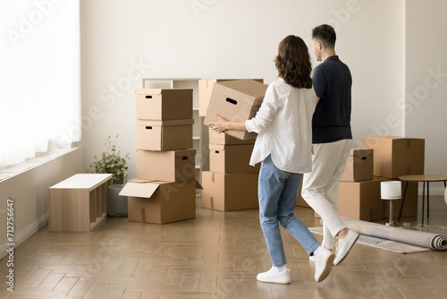 Young couple moving into new apartment after successful mortgage, carrying cardboard packages, walking in empty flat to heap of stacked boxes. Rear view of new homeowners photo