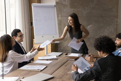 Happy young project manager woman offering plan, management strategy to diverse team, giving paper marketing reports to colleagues, smiling. Business coach training employees for sales growth