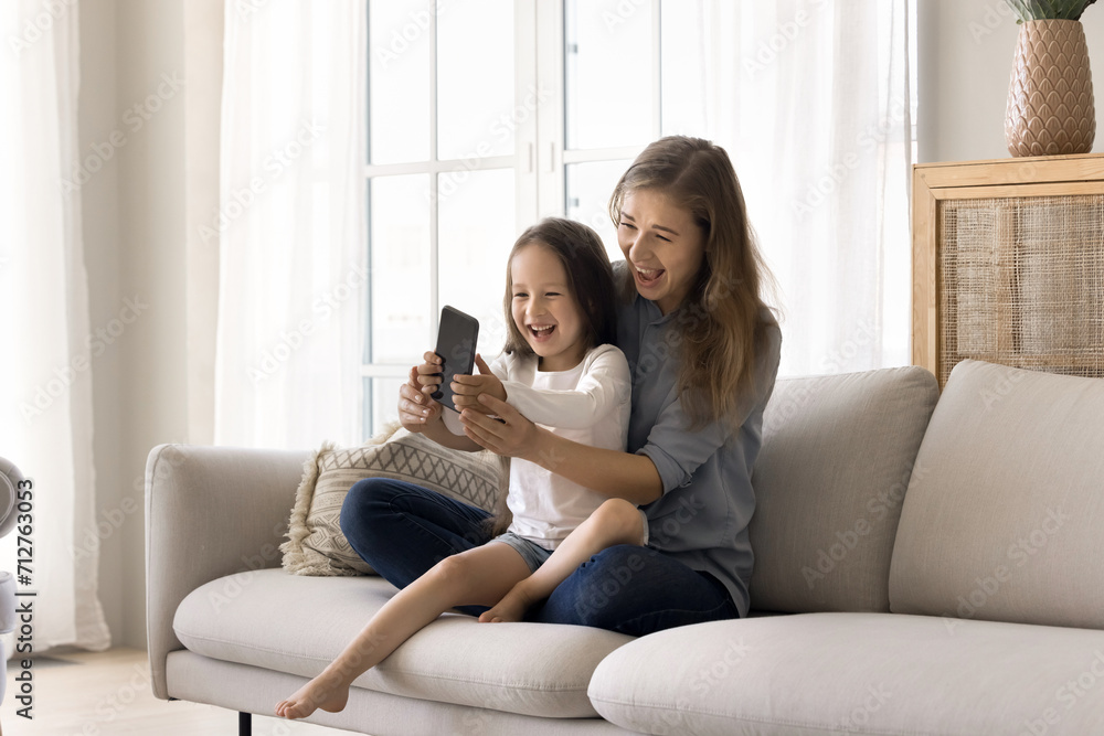 Overjoyed family relax sitting on sofa in living room, loving mother show funny mobile app to little daughter, pose for selfie photo, take video call, play new videogame, make video call to relatives Overjoyed family relax sitting on sofa in living room, loving mother show funny mobile app to little daughter, pose for selfie photo, take video call, play new videogame, make video call to relatives