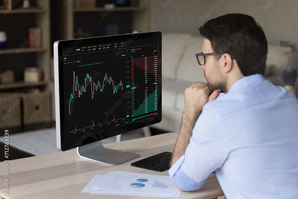 Serious man trader, analyst make stock market research, looks at pc screen learn information in charts and graphs, screen view with digital data over male shoulder. Crypto-currency, trading, analytics
