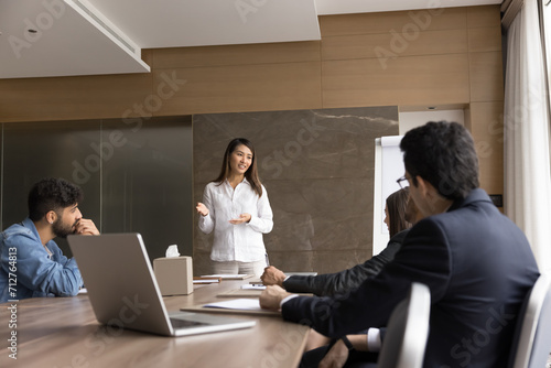 Positive Asian project manager woman presenting marketing ideas for startup to coworkers, partners. Young female leader standing at meeting table, talking to diverse team of colleagues