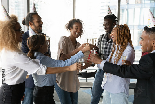 Raising glasses to success. Overjoyed diverse biracial colleagues have fun celebrate reward for project at company party in office. Excited mixed race business team toast greet leader with achievement