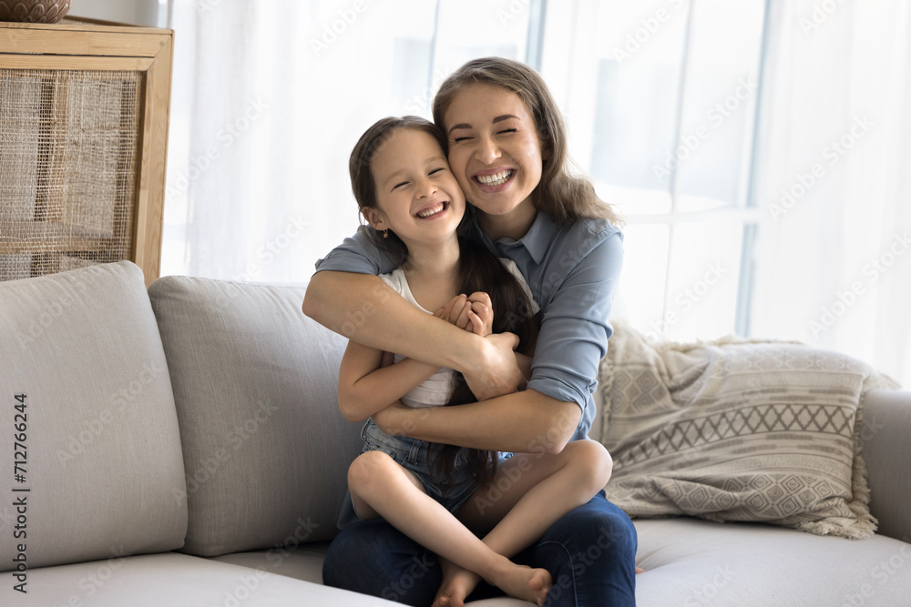 Loving mother sit on sofa cuddling tightly her little daughter, feeling pure love to child, spend priceless time together enjoy pastime at home. Cute girl sit on caring moms laps laugh look at camera Loving mother sit on sofa cuddling tightly her little daughter, feeling pure love to child, spend priceless time together enjoy pastime at home. Cute girl sit on caring moms laps laugh look at camera