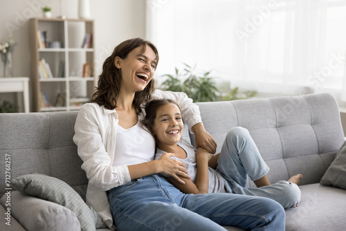Happy excited young mom and cheerful daughter kid having fun on soft couch at home, relaxing on sofa, looking away, laughing, enjoying family leisure, comfort, relaxation, close relationships