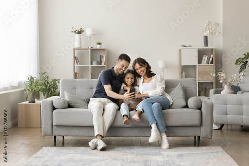 Positive young mom, dad and sweet girl kid taking family selfie on cozy comfortable couch, having fun with digital gadget in cozy stylish home interior, using smartphone for selfie together