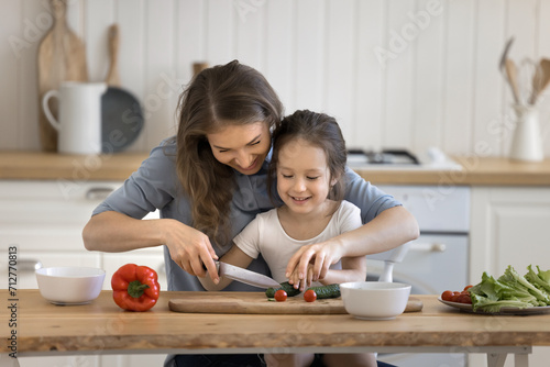 Loving mother teach to cook her little adorable daughter, holds knife, cutting fresh natural vegetables for dish, sit at table in modern cozy kitchen. Upbringing, kid development, share family recipe