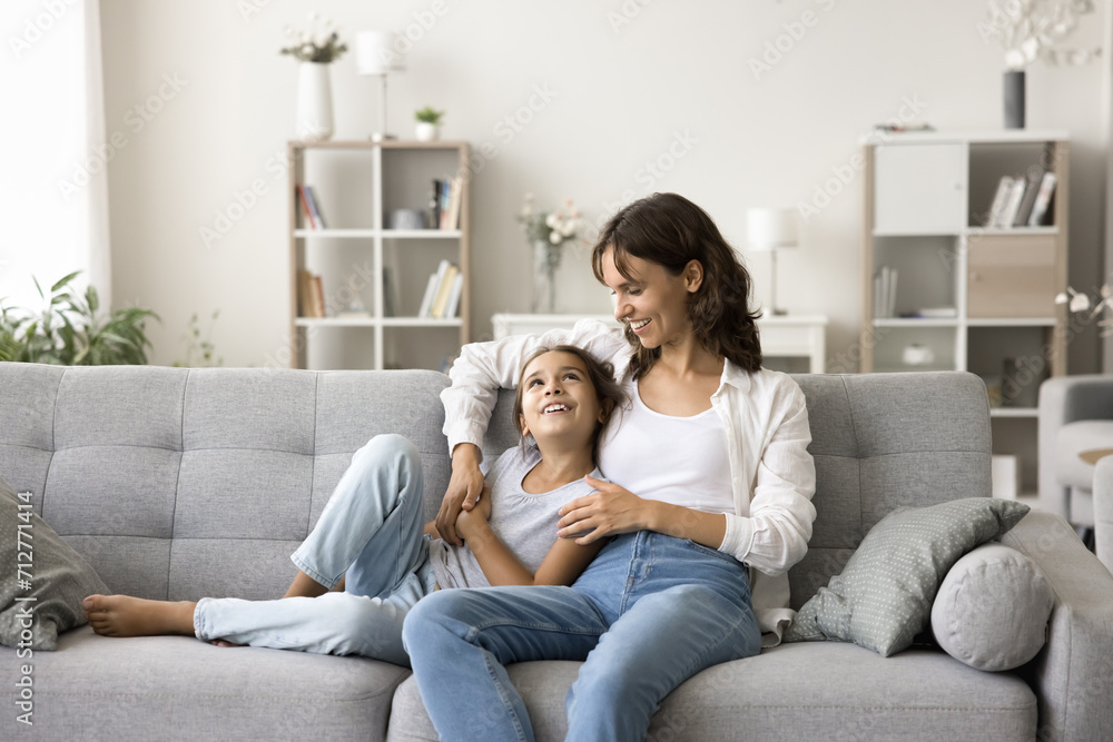 Happy young mom and cute little daughter resting on soft couch, hugging, chatting, enjoying family closeness, relationships, homey leisure time, talking, smiling, relaxing