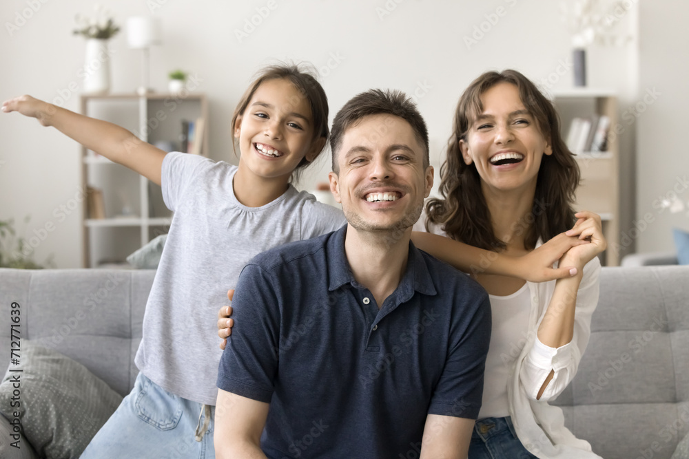 Cheerful happy parents and kid with open flying arms having fun on home couch, looking at camera with toothy smiles, enjoying family leisure, together, laughing. Video call screen, head shot portrait Cheerful happy parents and kid with open flying arms having fun on home couch, looking at camera with toothy smiles, enjoying family leisure, together, laughing. Video call screen, head shot portrait
