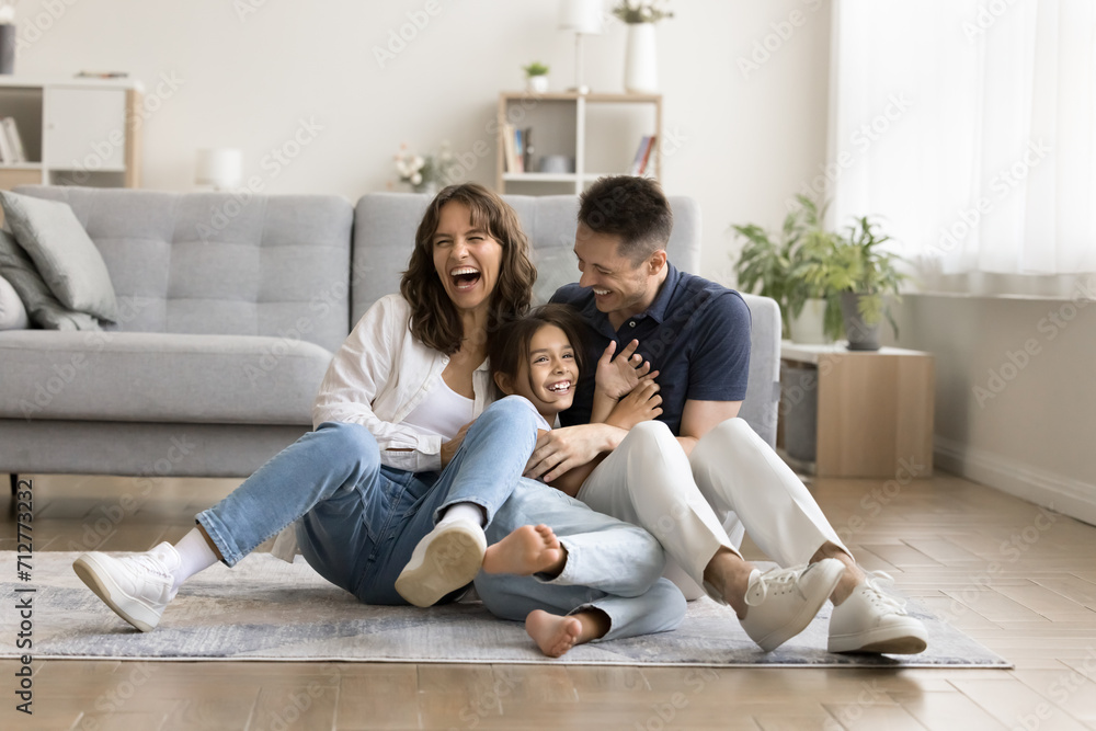 Cheerful excited parents tickling beloved kid girl, playing with child on heating floor in modern home interior, sitting on carpet, laughing, shouting for joy, enjoying family leisure Cheerful excited parents tickling beloved kid girl, playing with child on heating floor in modern home interior, sitting on carpet, laughing, shouting for joy, enjoying family leisure