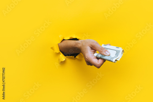 A female hand appears in the hole in torn yellow paper and squeezes two hundred-dollar bills. Concept of poverty, benefits, scholarships, and stinginess. Isolated. Copy space. photo