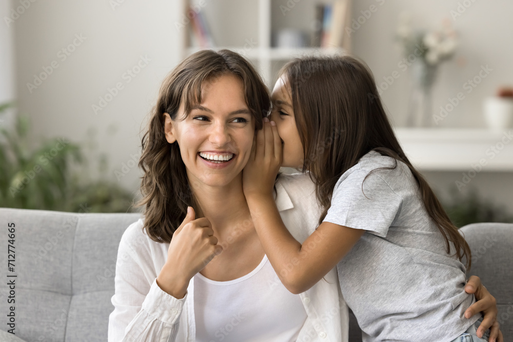 Cheerful positive little girl whispering secret at laughing mothers ear. Happy mom and daughter kid having fun on home sofa, talking, laughing, feeling closeness, trust, enjoying family leisure Cheerful positive little girl whispering secret at laughing mothers ear. Happy mom and daughter kid having fun on home sofa, talking, laughing, feeling closeness, trust, enjoying family leisure
