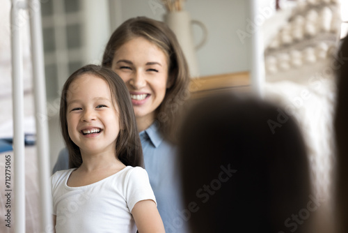 Happy mother and cute little daughter with pretty appearance looking in mirror, do morning routine, enjoy grooming process, loving parent teach her child to self-care, spend leisure together at home photo
