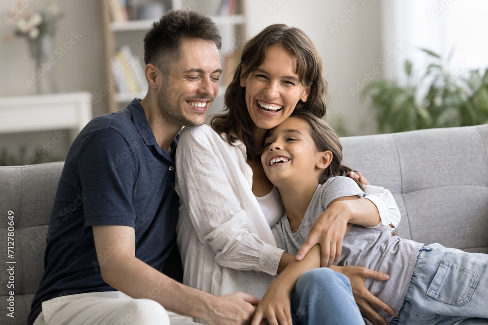 Excited happy young mother and father hugging little daughter kid on home sofa, tightening child with love, care, smiling, laughing, looking at camera, enjoying parenthood, leisure Excited happy young mother and father hugging little daughter kid on home sofa, tightening child with love, care, smiling, laughing, looking at camera, enjoying parenthood, leisure
