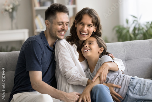 Excited happy young mother and father hugging little daughter kid on home sofa, tightening child with love, care, smiling, laughing, looking at camera, enjoying parenthood, leisure