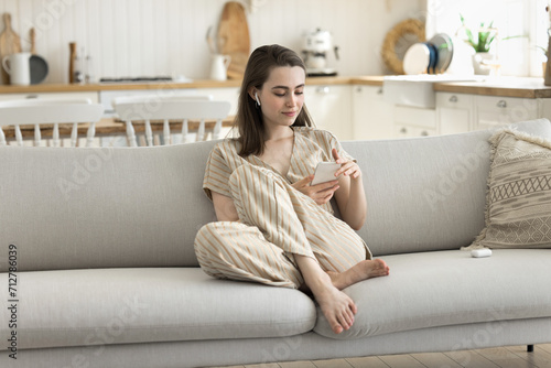 Young woman relaxing on cozy couch with modern smartphones, wear wireless earphones listen received audio messages from friend, choose playlist, music using subscription services, play online games