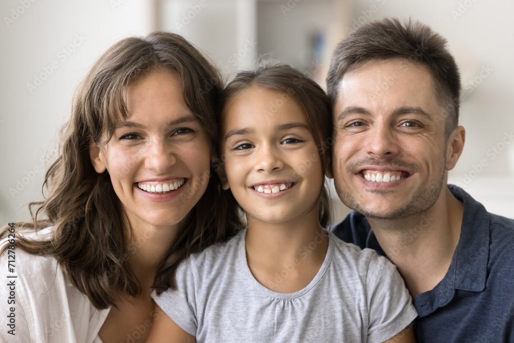 Happy beautiful young mom, handsome dad, cute daughter girl posing together with heads touch and toothy smiles, looking at camera, laughing. Parents and child home head shot portrait Happy beautiful young mom, handsome dad, cute daughter girl posing together with heads touch and toothy smiles, looking at camera, laughing. Parents and child home head shot portrait