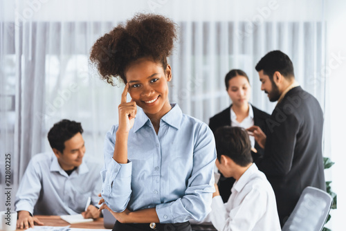 Young African businesswoman poses confidently with diverse coworkers in busy meeting room background. Multicultural team works together for business success. Office lady portrait. Concord