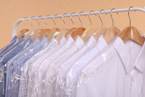 Dry-cleaning service. Many different clothes in plastic bags hanging on rack against beige background, closeup