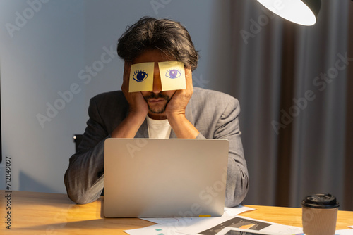 Sleeping clerk hides eyes with sticky notes, open eyes drawn on adhesive papers, he wants to sleep at workplace due lack of energy, chronic fatigue.