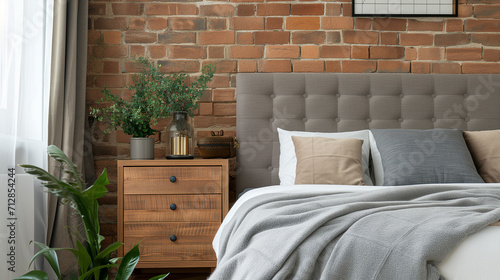 Wooden drawer nightstand near bed with grey fabric headboard. Loft interior design of modern bedroom with brown brick wall