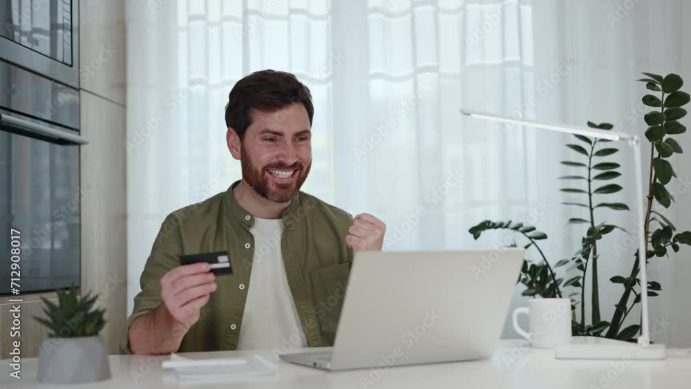 Joyful bearded man with credit card in hand making yes gesture while reading great news on modern laptop. Caucasian freelancer getting salary notification on email while sitting at home office. Joyful bearded man with credit card in hand making yes gesture while reading great news on modern laptop. Caucasian freelancer getting salary notification on email while sitting at home office.