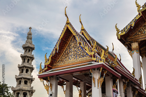 Fototapeta Naklejka Na Ścianę i Meble -  Decorated roof at Wat Suthat in Bangkok, Thailand. Intricate colored and golden ornamentation. Buildings and status on either side. Blue sky and clouds behind. 
