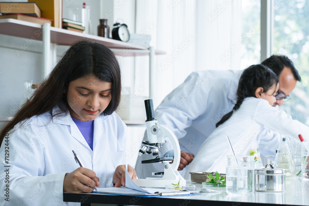 Indian scientist woman student using microscope do experiment and record results of experiment at biology class in school laboratory while teacher teaching kid student. Education, science concept Indian scientist woman student using microscope do experiment and record results of experiment at biology class in school laboratory while teacher teaching kid student. Education, science concept