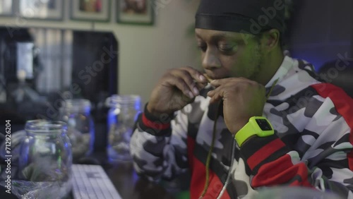 side view of a black man with a black head wrap is licking a blunt to seal the marijuana into the tobacco wrapper with mason jars of cannabis buds photo