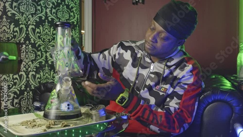 wide shot of a black man picking up a bag of marijuana bud and smelling the aroma before breaking the bud up into the marijuana grinder with a glass water bong on the table photo