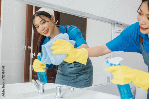 Maid with rubber gloves wipes bathroom mirror ensuring shiny reflection with a rag. Hotel service highlights professional cleaning ensuring purity and hygiene in modern settings. Maid cleaning at home