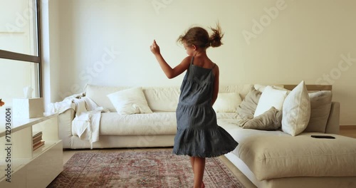 Cute little girl in casual summer dress sings a song and dancing in modern living room, lively moving, jumping, looking happy and untroubled. Funny adorable preschooler child having fun alone at home photo