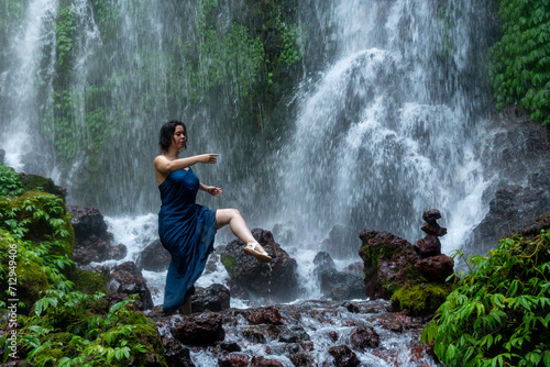 Cinematic portrait of the young beautiful woman standing in front of a waterfall in an blue dress.