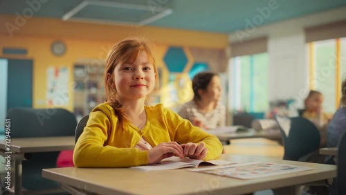 Portrait of a Cute Smiling Redhead Girl Sitting Behind a Desk in Class in Elementary School. Young Pupil is Looking at Camera, Smiling. Kids Educated in Modern Primary School photo