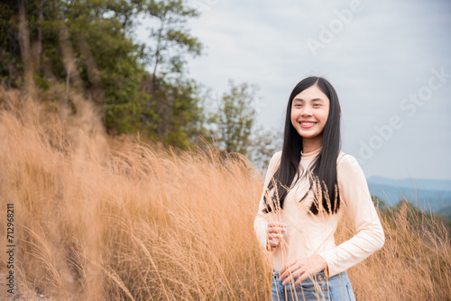 Beautiful Asian girl in casual walking in the field with golden color flower grasses in sunlight, lifestyle adorable young woman enjoy smiling feeling freedom happy in autumn season at sunset