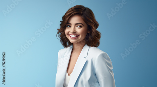 A radiant young woman in a chic white blazer poses with a confident smile against a soft blue background.