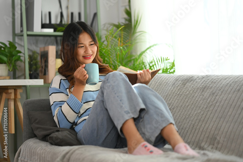 Relaxed young woman reading book and drinking coffee on couch at home