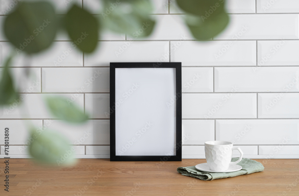 A black frame with a blank canvas against a white tile wall and on a wooden tabletop