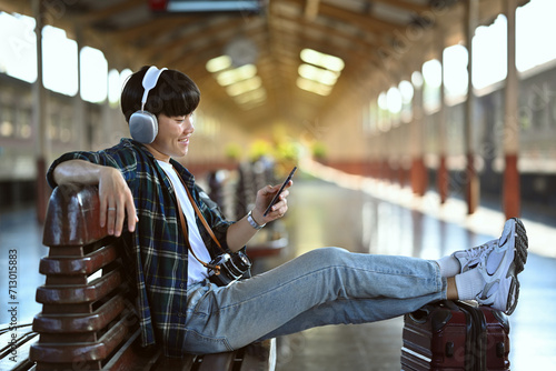 Happy young man in headphone using mobile phone during waiting for train at railway station