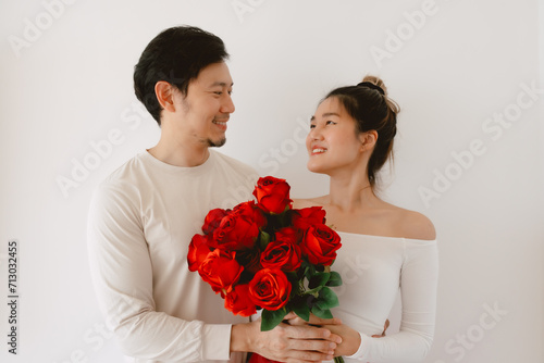 Happy Asian couples holding red roses, both woman and man smiling looking to each other, enjoy good time together on Valentines, isolated over white wall.