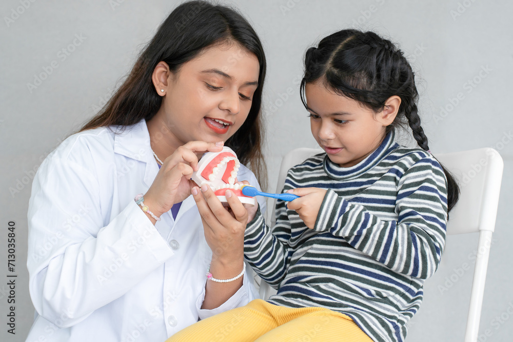 Cute Asian little girl learning how to brush her teeth and gums on a jaw model from young Indian dentist with toothbrush at dentist's clinic. Oral and Dental Health Cute Asian little girl learning how to brush her teeth and gums on a jaw model from young Indian dentist with toothbrush at dentist's clinic. Oral and Dental Health