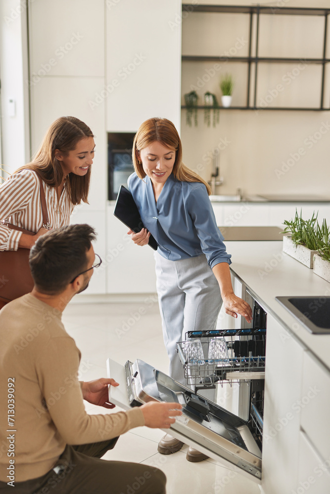Female shop assistant helping young couple choose new dishwasher Female shop assistant helping young couple choose new dishwasher