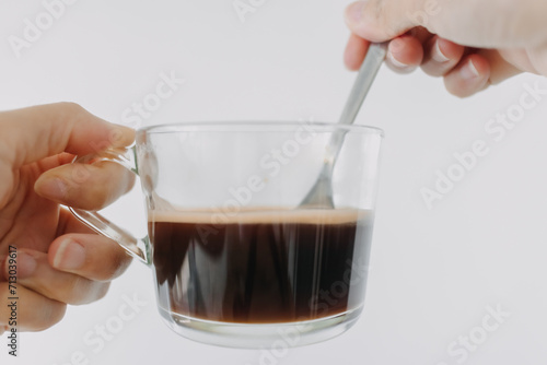 Close up of woman hand holding hot americano coffee clear mug with tea spoon, drink isolated over white background wall.
